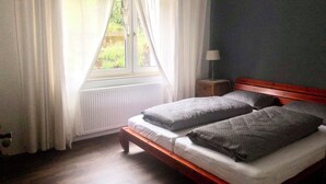 Una camera da letto con un letto in legno, tende bianche, una finestra vista verde e un comodino con una lampada.