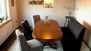 Zona pranzo con tavolo in legno, sedie imbottite nere e grigie, lampadario e una piccola mensola con oggetti decorativi.