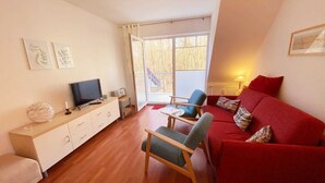 A living room with a red sofa, a TV on a wooden cabinet, a chair, and a large window with a view of trees. - Vacation apartment Haus Küste 9 (Graal-Müritz)