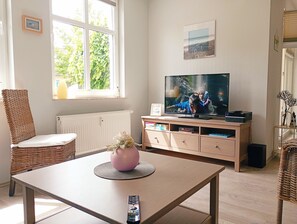 A living room with a television, a wicker chair, a table with a vase, and a window with greenery outside.