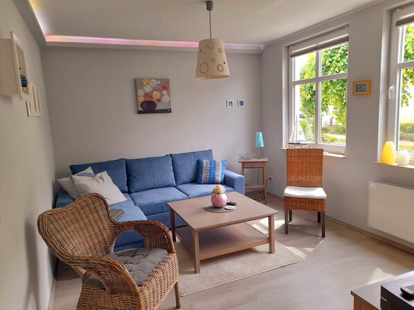 A living room with a blue sofa, wicker chairs, a wooden coffee table, and a large window with greenery outside.