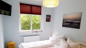 A bedroom with a bed, a window with blinds, a radiator, a small stool, and a painting on the wall. - Vacation apartment Seeadler 1 (Graal-Müritz)