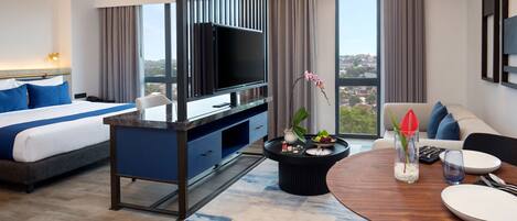 Una habitación de hotel moderna con una cama, un televisor, un comedor con mesa y sillas, y vistas a la ciudad a través de grandes ventanales.
