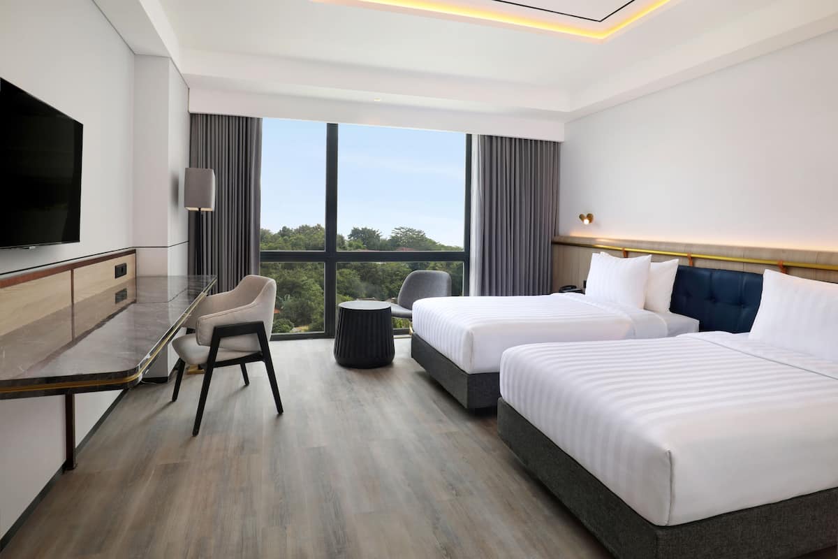 A hotel room with two beds, a desk, a chair, a TV, and a large window with a view of trees.