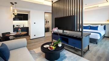 Een moderne hotelkamer met een bank, een eettafel met een schaal fruit, een televisie, een bed met blauw beddengoed en een bureau met een computer.