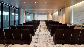 Un salón de conferencias con filas de sillas orientadas hacia una pantalla de proyección, ventanas amplias y una pared de mármol.