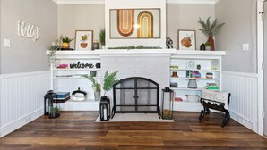 A living room with a fireplace, shelves with books and decorative items, and a large framed artwork above the mantel. - Cute Cottage close in Downtown Waco close to Magnolia, Baylor Uni., Cameron Park (Waco)