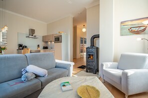 A modern living room with a sofa, armchair, and a table with a book and decorative item. A kitchen with appliances and a stove is visible in the background. - Villa Seestern, apt. 3 (Heringsdorf)