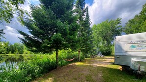 A campsite with a large tree, a hammock, picnic tables, and a water body.