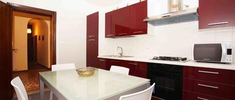 Une cuisine compacte avec une table à plateau en verre, des chaises blanches et des placards rouges.