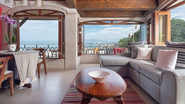 Un espacio de sala acogedor con un sofá, una mesa de centro de madera y una mesa de comedor con sillas. Amplias ventanas ofrecen vistas al mar.