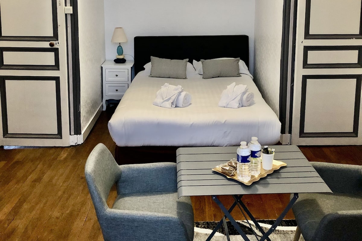 Une chambre d’hôtel avec un lit bien fait, deux tables de chevet, une petite table sur laquelle se trouve un plateau avec divers objets, et deux chaises.