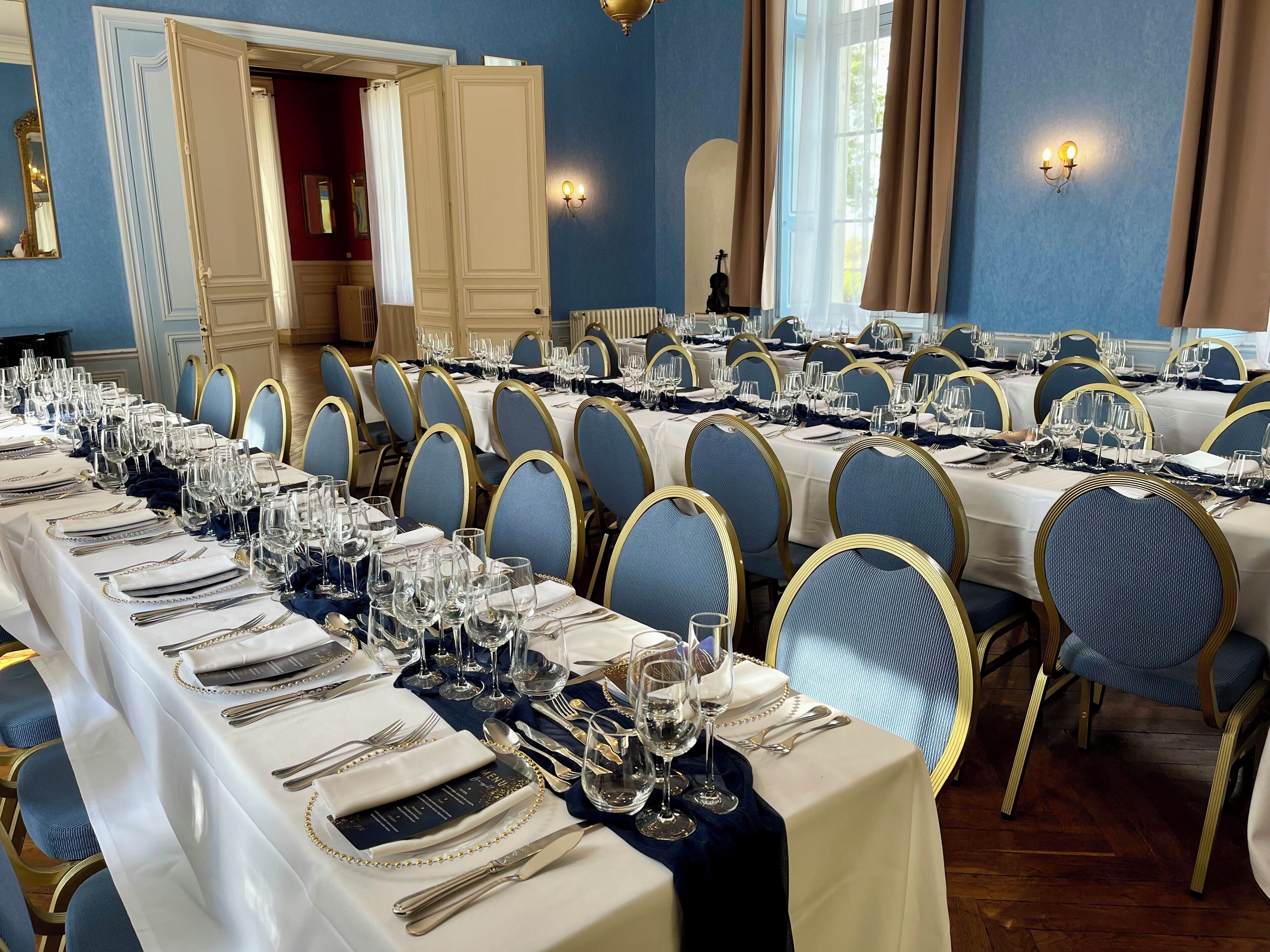 A formal dining room with long tables set for a meal, blue chairs, and a blue and gold color scheme.