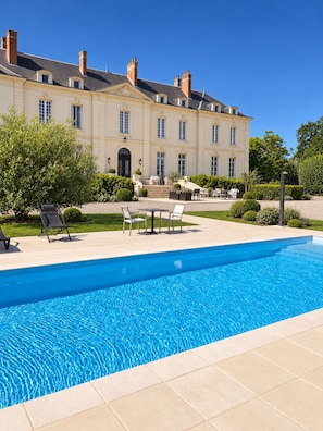 Ein Pool mit Liegestühlen und einer Terrasse vor einem großen Gebäude mit vielen Fenstern und einem zentralen Eingang.
