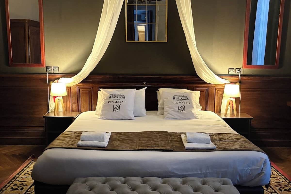 Une chambre d’hôtel avec un grand lit, deux tables de chevet, un lit à baldaquin, un miroir et un tapis à motifs.
