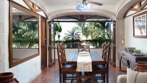 A dining area with a wooden table and chairs, a ceiling fan, and a view of palm trees through open doors.