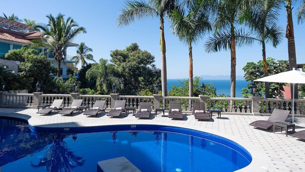 A pool area with lounge chairs, a thatched roof structure, and a view of the sea.
