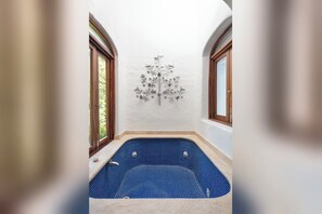 Zona de spa con un jacuzzi azulado de azulejos, paredes blancas y una escultura decorativa de un árbol metálico.