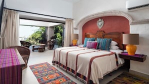 A bedroom with a bed, a table, chairs, a lamp, and a view of greenery outside. - Rent the Entire Floor at Luxury Resort: 5th Floor Suites Spectacular Ocean Views (Puerto Vallarta)