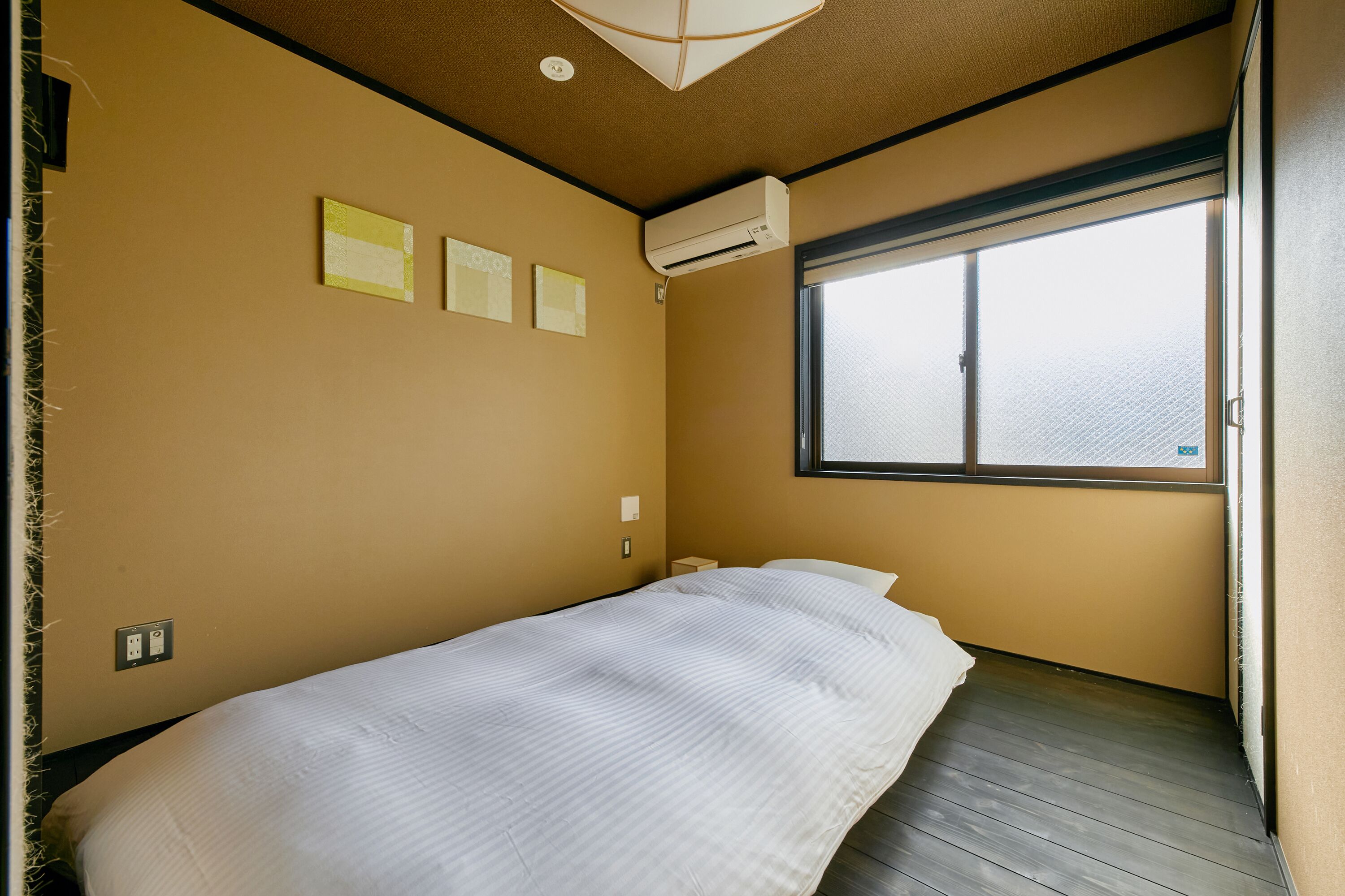 A bedroom with a single bed, an air conditioning unit, a window, and a wall-mounted electrical outlet.