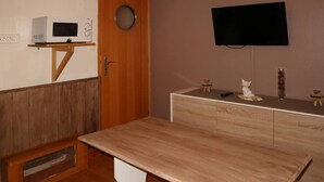 Une pièce de taille modeste, avec une table en bois, un téléviseur fixé au mur, un four à micro-ondes posé sur une étagère et un petit meuble.
