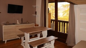 Une pièce avec une table et des bancs en bois, un téléviseur fixé au mur et une fenêtre donnant sur des arbres.