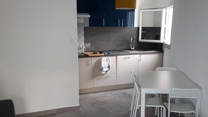 Une cuisine moderne avec des meubles de couleur bleu foncé, un plan de travail blanc et un luminaire suspendu jaune. On y trouve un coin repas avec une table et des chaises blanches, ainsi qu’un tapis à rayures sur le sol.