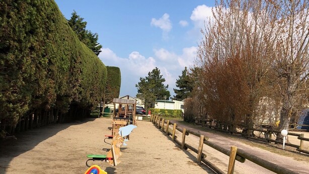 A playground with various play equipment, including a swing set and a seesaw, surrounded by trees and a wooden fence. - Mobil Home 3 chambres (Port-Bail-sur-Mer)
