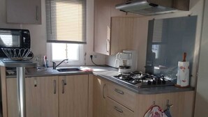 A compact kitchen with a microwave, sink, stove, and wooden cabinets. - Mobil Home 3 chambres (Port-Bail-sur-Mer)