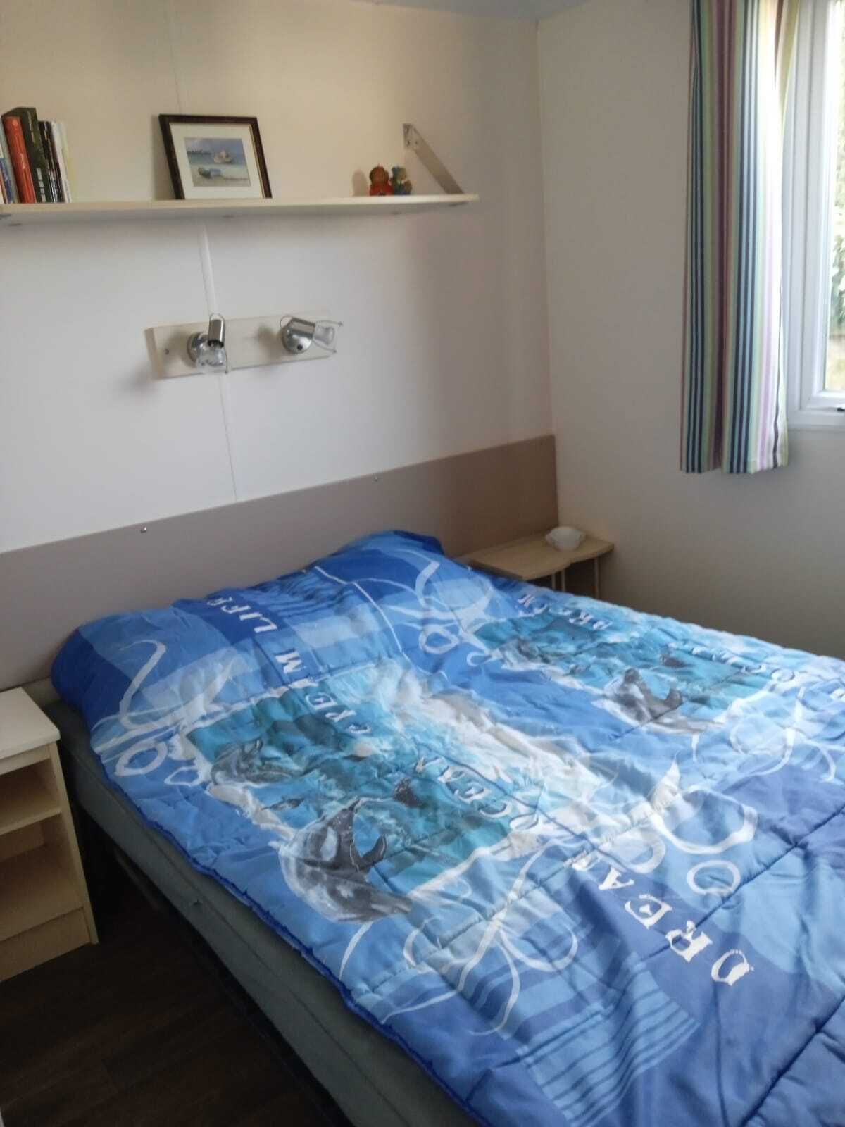 A bedroom with a bed, a nightstand, a shelf with books and a picture, and a window with curtains.