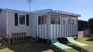 A mobile home with a striped awning, a small porch, and two green-striped lounge chairs. - Mobil Home 3 chambres (Port-Bail-sur-Mer)