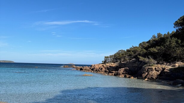 Ein Strand mit klarem, blauem Wasser, sandigem Ufer und felsigen Klippen mit Bäumen.