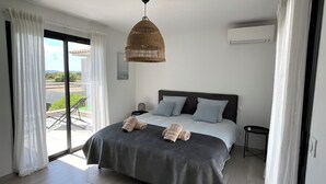 Ein Schlafzimmer mit Bett, einer Pendelleuchte, einem Nachttisch und Blick ins Freie durch eine Schiebetür.