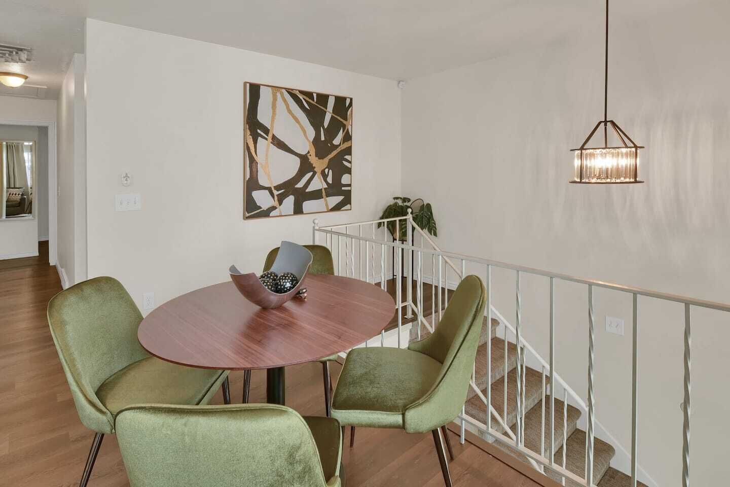 A dining area with a round table, two green chairs, a bowl with decorative items, a modern abstract painting, and a hanging light fixture.
