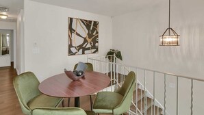 A dining area with a round table, two green chairs, a bowl with decorative items, a modern abstract painting, and a hanging light fixture.