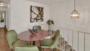 A dining area with a round table, two green chairs, a bowl with decorative items, a modern abstract painting, and a hanging light fixture.