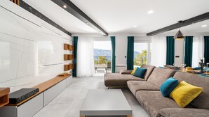 Una sala de estar moderna con sofá, mesa de centro y televisor integrado. Hay un balcón con vista a las montañas.