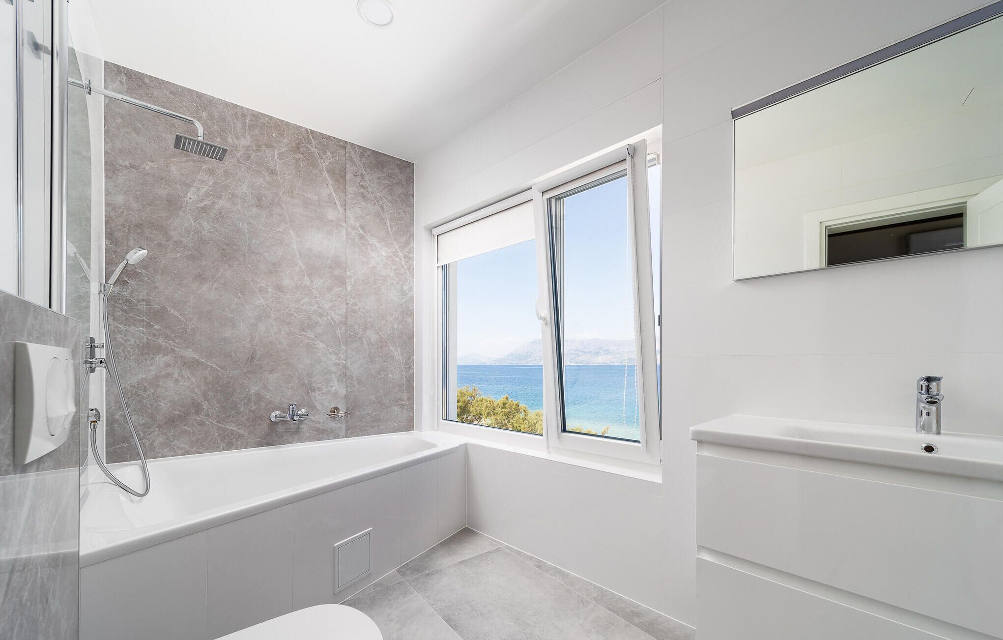 A modern bathroom with a bathtub, a shower, and a sink. There is a window providing a view of the sea.