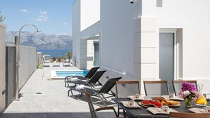 Un área de piscina moderna al aire libre con sillones de descanso y una mesa de comedor con alimentos para el desayuno.