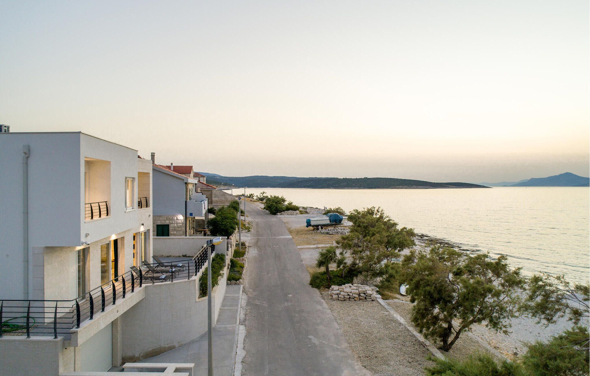 A coastal road with modern buildings, a clear view of the sea, and a distant mountain range.