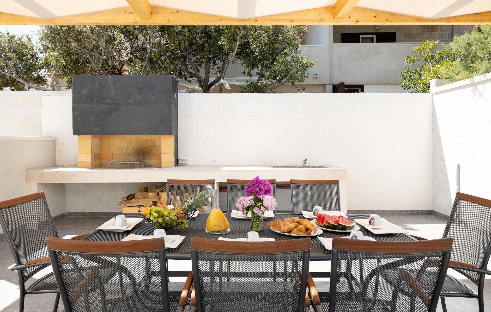 Un espace extérieur aménagé pour les repas, avec un barbecue, des sièges et une table décorée de fruits, de fleurs et de pâtisseries.
