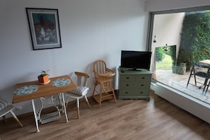Une pièce comprenant une table à manger en bois, deux chaises, un téléviseur posé sur un meuble et une fenêtre donnant sur un paysage verdoyant.