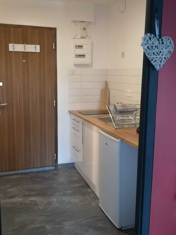 A compact kitchen with a wooden door, white tiled backsplash, a small sink, and a compact refrigerator.