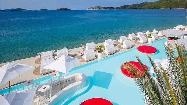 A resort pool with white lounge chairs and umbrellas, surrounded by a clear blue sea and distant green hills.