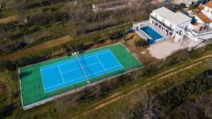 An aerial view of a tennis court adjacent to a modern house with a swimming pool.