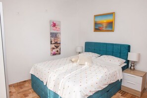 Une chambre à coucher avec un lit recouvert d’un tissu turquoise, une literie blanche ornée de motifs colorés, une table de chevet avec une lampe et un tableau représentant un coucher de soleil, accroché au mur.
