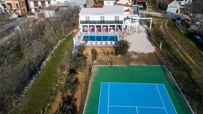Vue aérienne d’un bâtiment moderne doté d’une piscine, d’un court de tennis et d’un espace de détente extérieur.