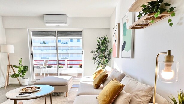 Un soggiorno moderno con un divano, un tavolino e una lampada da terra.  Un balcone offre vista su edifici e un vaso con una pianta sul davanzale.