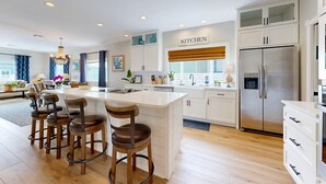 A modern kitchen with a central island, bar stools, a stainless steel refrigerator, and white cabinetry. - Shared Pool, Boardwalk to Beach and Golf Cart Included (Port Aransas)