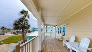A wooden deck with white Adirondack chairs and a view of a residential area with palm trees and houses. - Shared Pool, Boardwalk to Beach and Golf Cart Included (Port Aransas)
